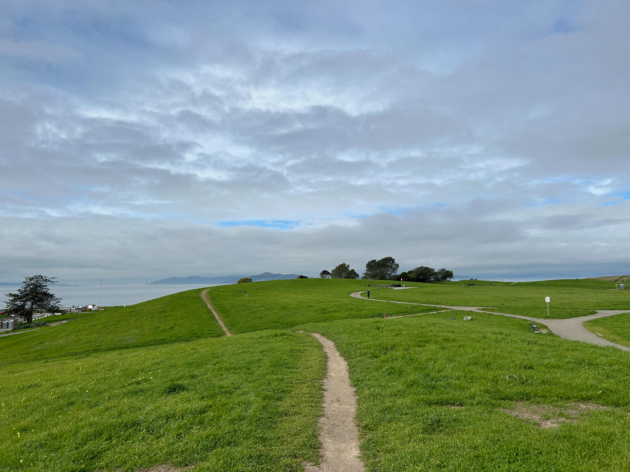 Grassy rolling hills with curving dirt paths under a cloudy sky, with a glimpse of blue and distant mountains by the water.